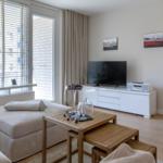 Wohnzimmer mit Sofa, TV-Schrank und Holztisch. Fenster mit Jalousien und Blick auf Balkon.