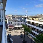Blick auf einen Platz mit Gebäuden und einem Turm unter blauem Himmel.
