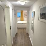 Flur mit Holzboden und weißen Wänden. Im Hintergrund ist ein Schlafzimmer mit Bett und Vorhang sichtbar.