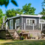 Wooden house with terrace, garden furniture, and barbecue area. Surrounded by trees under blue sky.