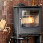 A wood stove with a burning fire, next to a basket of firewood and a red bucket.
