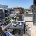 Terrasse mit Tisch, Stühlen und Blumen. Blick auf Gebäude und Meer.