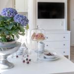Tisch mit Blumen, Donuts und Kirschen. Hintergrund: Fernseher und Schrank.