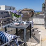 Terrasse mit Tisch, Stühlen und Blumen. Blick auf Gebäude und Meer.