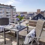 Terrasse mit Tisch, zwei Stühlen und Blumen. Blick auf Wohngebiet unter blauem Himmel.