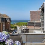 Blick von Balkon auf Straße, Häuser und Meer unter blauem Himmel mit Hortensien im Vordergrund.