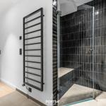 Dusche mit Glaswand und schwarzen Fliesen. Heizkörper an der Wand. Holzboden im Flur.