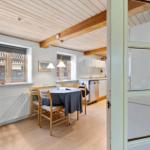 Dining room with table, chairs, and kitchen area. Wooden ceiling with beams and windows.