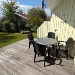 Terrasse mit Tisch und Stühlen neben gelbem Haus. Grünflächen und Wasser im Hintergrund.