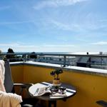 Terrasse mit Blick auf das Meer und Gebäude. Tisch, Stuhl, Pflanze und Zeitschrift.