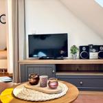 TV auf Holzboard mit Lautsprechern und Pflanze. Runder Tisch mit Kerzen und Korb. Sicht auf Küche.