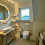Badezimmer mit runden Spiegel, Waschtisch und Toilette. Fenster mit Blick auf Landschaft.