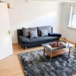 Ein Wohnzimmer mit grauem Sofa, Glas-Tisch und Holzboden. Fenster mit weißen Vorhängen.