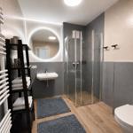 Badezimmer mit Dusche, Waschbecken und Toilette. Holzboden und graue Wandfliesen.