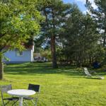Grüner Garten mit Tisch, Stühlen und Liegestuhl. Im Hintergrund ein Haus mit rotem Dach.