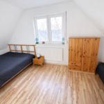 Schlafzimmer mit zwei Betten, Holzschrank und Fenster. Holzfußboden und weiße Wände.