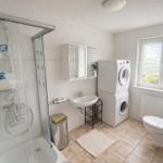 Badezimmer mit Dusche, Waschmaschine, Toilette und Fenster.