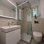 Badezimmer mit Dusche, Waschbecken und WC. Spiegel mit Beleuchtung und Fenster.
