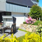Terrasse mit Tisch, Stühlen und Liege unter blauem Himmel. Blühender Garten mit Steinmauer.