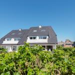 Modernes Haus mit dunklem Dach und weißen Fenstern, umgeben von grünen Sträuchern unter blauem Himmel.
