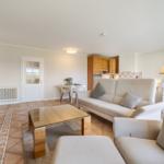 Living area with sofa, armchair, glass table, and kitchen area in the background.