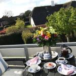 Terrasse mit Tisch, Stühlen und Blumenstrauß. Blick auf grüne Hecken und Häuser mit Strohdächern.