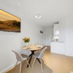 Dining area with table and chairs, kitchen in background, wall art and wooden floor.