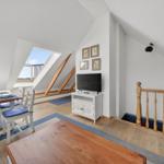 Dachgeschosswohnung mit Essbereich, TV und Balkonzugang. Holzdielen und weiße Wände.
