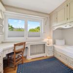 Schlafzimmer mit Schreibtisch, Bett und Fenster. Holzfußboden und blaues Teppichmuster.