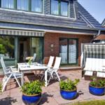 Terrasse mit weißem Tisch, Stühlen und Strandkorb. Blaue Töpfe mit Pflanzen. Haus mit Glasfront und Ziegelfassade.