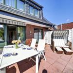Terrasse mit Tisch, Stühlen und Strandkorb. Haus mit Glasfront und rotem Ziegelmauerwerk.