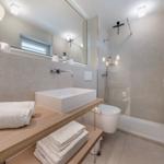 Badezimmer mit Dusche, Waschbecken und Toilette. Weiße Handtücher auf Holzregal.