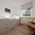 Schlafzimmer mit Bett, Schubladen, Tisch und Stuhl. Fenster mit Blumen und Bild an der Wand.