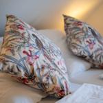 Zwei Kissen mit floraler Vogelmusterung auf einem Bett mit weißer Bettwäsche.