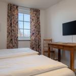 Schlafzimmer mit Bett, Schreibtisch, Stuhl und Wandbild. Fenster mit floralen Vorhängen.