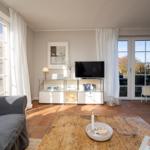 Wohnzimmer mit TV, Holztisch und großen Fenstern. Terrakottaboden und weiße Vorhänge.