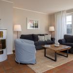 Wohnzimmer mit Sofa, Sessel, Kamin und Holztisch. Terrakottaboden und Fenster mit Vorhängen.