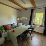 Ein Raum mit Tisch, Stühlen und Sofa. Holzbalken an der Decke und Fenster mit Blick auf Bäume.