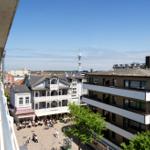 Blick auf eine belebte Straße mit Gebäuden und einem Turm im Hintergrund.