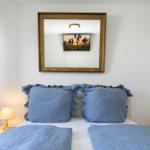 Doppelbett mit blauen Kissen und Decken, zwei Nachttische mit Lampen, Wandspiegel mit Bild.