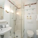 Badezimmer mit Dusche, Waschbecken und Toilette. Weiße Fliesen und Spiegel.