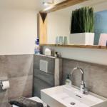 Badezimmer mit Waschbecken, Spiegel und Toilette. Holzrahmen und Pflanze sichtbar.