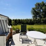 Terrasse mit Liegestuhl, Tisch und Strandkorb. Grünfläche und Bäume im Hintergrund.