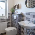 Badezimmer mit Toilette, Waschbecken und Fenster. Wandverkleidung aus grauen Fliesen.