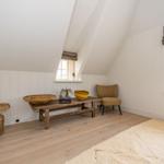 Ein Zimmer mit schrägen Wänden, Holzboden und zwei Fenstern. Möbel aus Holz und Rattan.