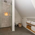 Dusche mit Glaswand, Toilette und Holzregal mit Handtüchern unter Dachschräge.