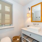Badezimmer mit Waschbecken, Spiegel, Toilette und Fenster mit Jalousie.