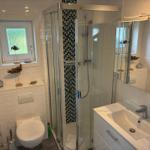 Badezimmer mit Dusche, Waschbecken und Toilette. Fenster mit Blick auf Grün.