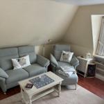 Ein Wohnzimmer mit grauem Sofa, Sessel und weißem Couchtisch. Fenster mit weißen Vorhängen.