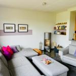 Wohnzimmer mit grauem Sofa, Kamin und Bücherregal. Küche im Hintergrund.